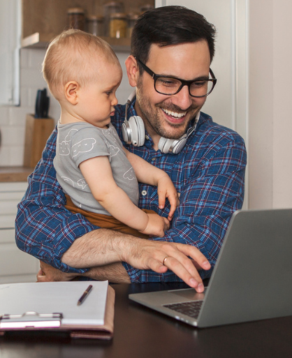 father holding his baby while trying to use his laptop
